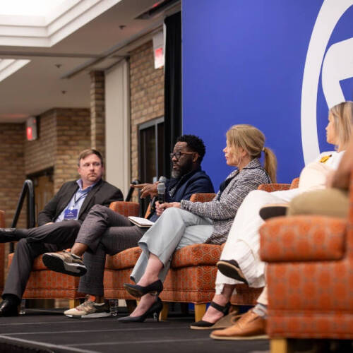 A group of people, both men and women are seated on orange patterned chairs on a stage, engaged in a panel discussion. The background features a large blue wall with a white logo, and the setting appears to be a conference or professional event.